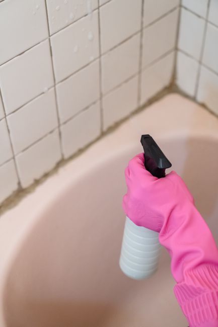Close-up of a hand wearing a bright pink rubber glove holding a white spray bottle with a black nozzle, aimed at the corner of a bathroom bathtub. The background features white ceramic tiles with visi