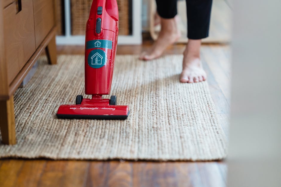 Close-up view of a vacuum cleaner with a transparent dust container filled with debris, connected to a flexible, patterned hose and a black floor nozzle, situated on a soft pink carpet in a well-lit r