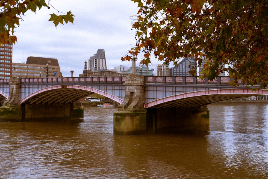 Photograph of Hammersmith Bridge spanning the river in a cityscape, with architectural details visible on the bridge's pink-painted iron structure supported by stone pillars. The river below shows gen